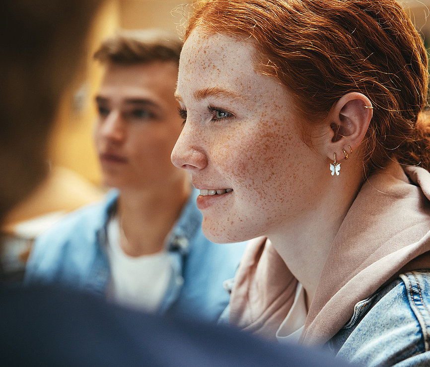 Foto eines jugendlichen Mädchens, welches interessiert in eine Runde Jugendlicher schaut